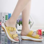 Sandales orthopédiques jaunes portées par une femme sur un carrelage avec livres et mur blanc