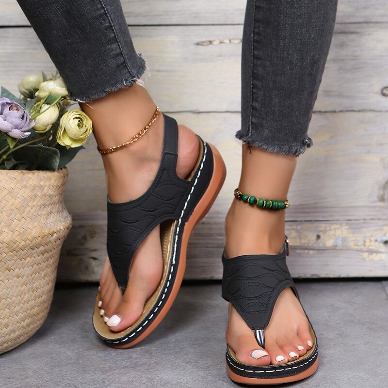Sandale orthopédique noire d'été pour un confort maximal pour femme, les sandales aux pieds d'une femme debout, on ne voit que ses pieds, devant un mur en bois.